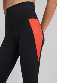 Czarne legginsy z wysokim stanem, z jasnopomarańczowym panelem po boku. Materiał jest gładki z widocznymi detalami szwów.