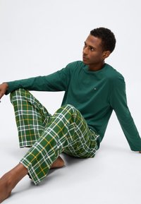 Πράσινο μακρυμάνικο πουκάμισο συνδυασμένο με πράσινες και λευκές πιτζάμες καρό με κίτρινες λεπτομέρειες. Το μοντέλο κάθεται, με γυμνά πόδια ορατά.