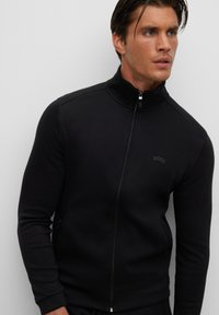 Svart zip-up sweatshirt med hög hals, tillverkad av slät tyg, med en subtil logotyp på bröstet och dragkedjefickor på sidorna.