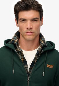 Joven con cabello oscuro que lleva una sudadera con cremallera de color verde oscuro sobre una camisa de cuadros y una camiseta blanca, expresión neutral, fondo claro y sencillo.