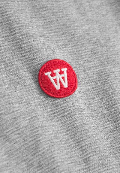 Tissu gris avec un patch circulaire rouge arborant des lettres "W" blanches brodées. La texture est douce et lisse. Le patch contraste avec le tissu.