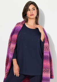 Marineblaues Oberteil mit Strassdetail "Perfect"; drapierter, farbenfroher Schal mit rosa, orange und lila Mustern; Model posiert mit subtiler Struktur in den Stoffen.