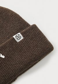 Brown knitted beanie with a ribbed texture. Features a white fabric label with a black logo on the cuff. Soft material and snug fit.