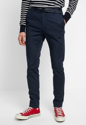 Uomo che indossa pantaloni slim fit blu navy, cintura nera, top a maniche lunghe a righe e sneakers alte rosse con lacci bianchi.