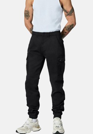 Mann trägt schwarze Cargohosen mit elastischen Bündchen, weiße Sneakers und ein weißes ärmelloses Shirt und zeigt Arm-Tattoos.