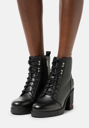 Bottes noires en cuir à hauteur de la cheville avec talons épais, lacets sur le devant, fermetures éclair latérales et œillets métalliques décoratifs portées sur des jambes à peau foncée.