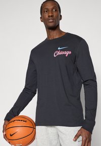 Schwarz langärmliges Shirt mit rotem und blauem "Chicago"-Grafik und Nike-Logo, kombiniert mit grauen Hosen und einem Basketball.