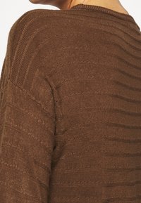 Inquadratura ravvicinata di una persona che indossa un maglione a maglia marrone con una sottile trama orizzontale e un colletto a coste.