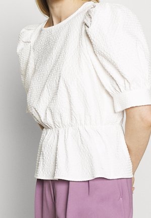 Vrouw in een getextureerde witte blouse met pofmouwen en een geplooide taille, gecombineerd met losse lavendelkleurige broek, tegen een effen achtergrond.