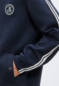 Ναυτικό μπλε μπουφάν με λευκές διπλές ρίγες στο μανίκι, χέρι σε τσέπη με κουμπί και κυκλικό έμβλημα λογότυπο FC Marc O’Polo Stockholm 1967.