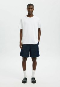 Vit bomull t-shirt med korta ärmar, tillsammans med mörkblå denimshorts. Modellen har på sig vita strumpor och svarta loafers.