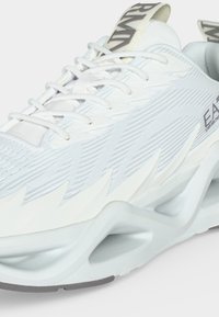 Gros plan sur une chaussure de sport blanche avec une tige texturée, un design géométrique de la semelle, des lacets et le logo EA7 Emporio Armani visible sur le côté et la languette du talon.