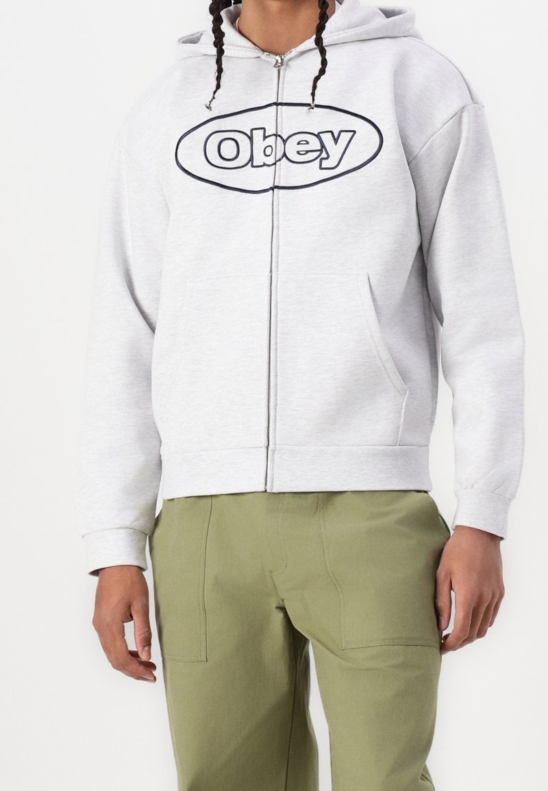 Personne portant un sweat à capuche zippé gris clair avec le logo "Obey" et un pantalon vert olive sur fond blanc uni.