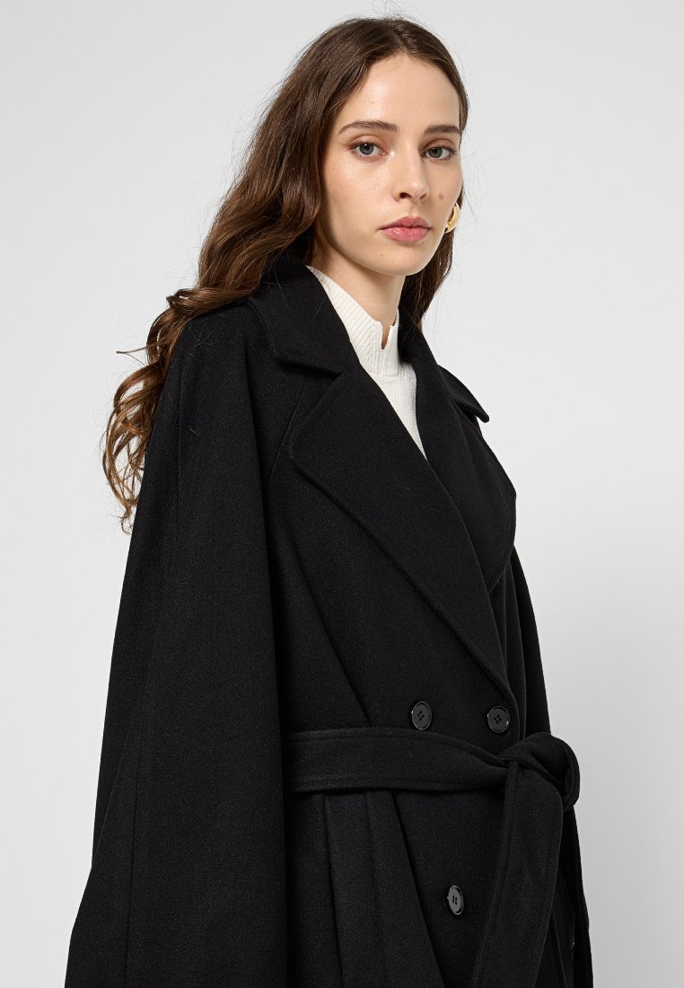 Czarny płaszcz z mieszanki wełny z podwójnym zapięciem, szerokimi klapami i wiązanym w pasie. Gładka tekstura z wyraźnie zarysowanym krojem.