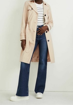 Kobieta ubrana w beżowy płaszcz trencz, białą pasiastą koszulę, ciemnoniebieskie dzwony i białe trampki, stojąca na jednolitym tle.