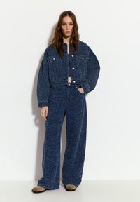 Džínová bunda a odpovídající široké kalhoty z texturované tmavě modré látky. Bunda má přední kapsy a knoflíky; kalhoty mají vysoký pas a volný střih.