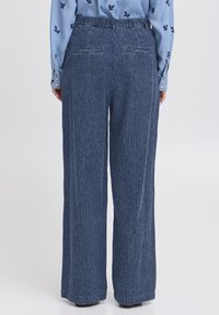 Persona que lleva pantalones de tiro alto y pierna ancha a rayas azul con una camisa de manga larga azul con pequeños patrones de hojas oscuras, de pie de espaldas.