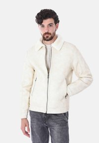 Giacca di shearling color crema con chiusura a zip, ampio colletto, tasche laterali e dettagli di texture sottili, indossata sopra jeans di denim scuri.