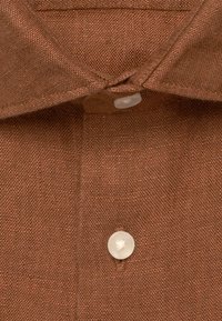 Gros plan sur le col texturé marron d’une chemise avec deux boutons blancs, un boutonné en haut et un en bas déboutonné.