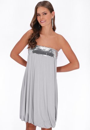 Vrouw in een zilverkleurige strapless jurk met pailletten op het lijfje en oorbellen in hartvorm, glimlachend met handen achter de rug op een witte achtergrond.