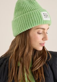 Zielona prążkowana dzianinowa czapka z odwiniętym rondem i białą etykietą z czarnym napisem. Miękka w dotyku, dobrze dopasowana i ciepła.