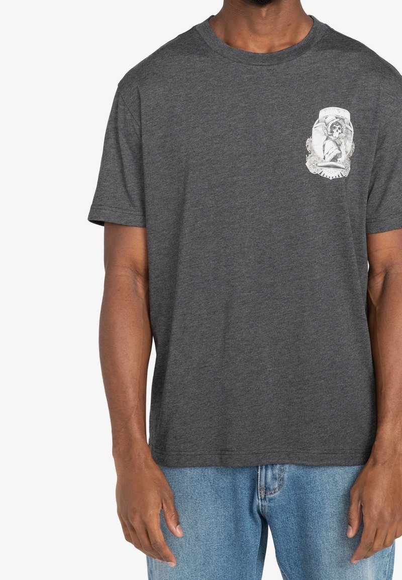 Mężczyzna ubrany w ciemnoszary T-shirt z krótkim rękawem z białą grafiką przedstawiającą postać szkieletu oraz jasnoniebieskie dżinsy, widoczny od szyi do bioder.