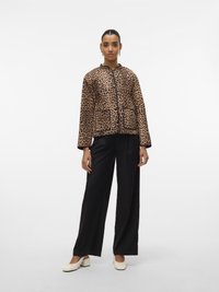 Woman wearing a leopard-print jacket with black trim, black wide-leg trousers, white block-heeled shoes, standing against a plain white background.