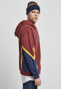 Bordeauxfarbener Hoodie mit marine- und gelben Akzenten, mit einem Logo auf der Vorderseite. Kombiniert mit einer marineblauen Mütze mit mehrfarbigen Streifen. Lässige Passform.