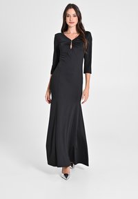 Schwarzes langärmliges Kleid mit figurbetontem Schnitt, gedrehtem Oberteil, Ausschnitt und bodenlangem Rock. Silbern hohe Absätze.