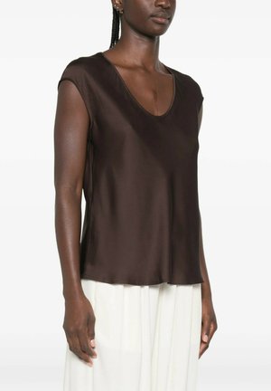 Donna che indossa un top marrone scuro senza maniche con scollo a V e vestibilità ampia abbinato a pantaloni bianchi a vita alta su uno sfondo semplice.