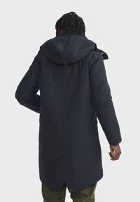 Long manteau imperméable bleu marine foncé avec capuche, dotée d'une fermeture éclair et d'une fermeture à pression, de poignets ajustables et d'une coupe droite.