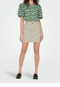 Blouse à manches courtes vert à motifs avec des manches bouffantes, associée à une jupe beige arrivant au genou et des mocassins noirs.