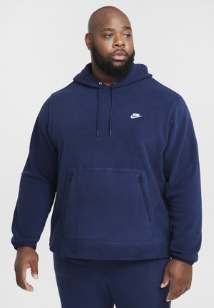 Marineblå Nike fleece hoodie med en kengurulomme, snoretræk i hætten og hvidt logo på brystet. Blød tekstur og afslappet pasform.