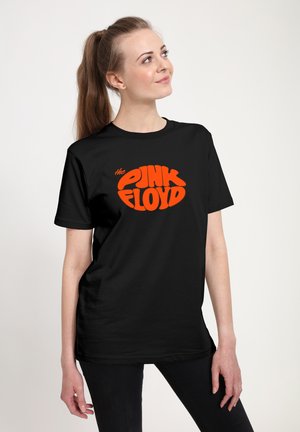 Jeune femme avec une queue de cheval porte un t-shirt noir avec un texte orange vif « The Pink Floyd », debout sur un fond clair uni.
