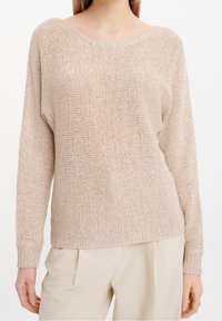 Femme portant un pull en maille beige à manches longues et encolure dégagée, associé à un pantalon crème clair.