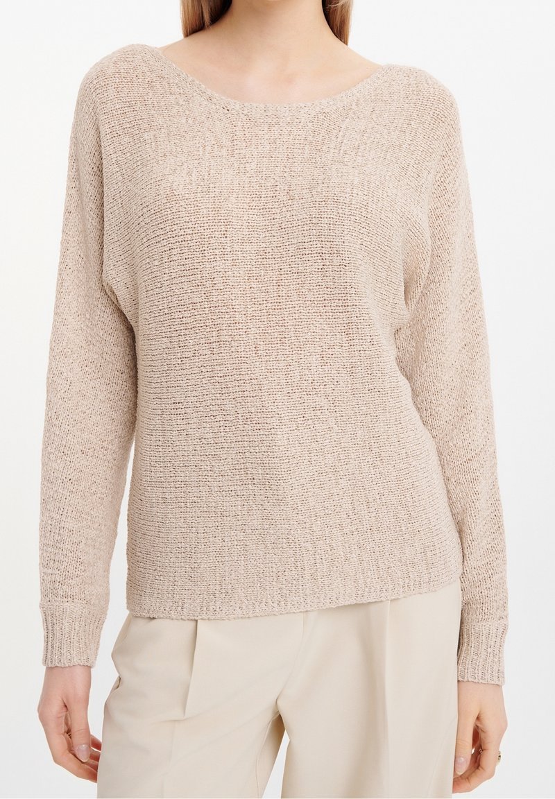 Femme portant un pull en maille beige à manches longues et encolure dégagée, associé à un pantalon crème clair.