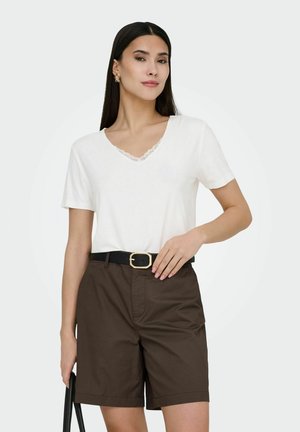 Woman with long dark hair wearing a white V-neck top with lace trim, brown shorts with black belt, holding a black bag.