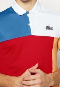 Mężczyzna w wielokolorowej koszulce polo z niebieskimi, białymi, czerwonymi i beżowymi częściami, z czarnym logo krokodyla na górnej prawej części klatki piersiowej.