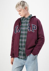 Bordeauxkleurige rits-hoodie met witte "GAP" letters, gedragen over een groen en marineblauw geruit hemdd; gecombineerd met lichtblauwe spijkerbroek en een casual outfit.