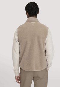 Beige Fleece-Weste mit weißen Ärmeln, Stehkragen und einem leicht geschwungenen Saum. Verfügt über ein Etikett am Kragen und gerippte Bündchen.