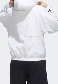 Hvid vindjakke med hætte, elastiske manchetter og kant. Har en glat stoftekstur og brandingdetalje på ærmet.