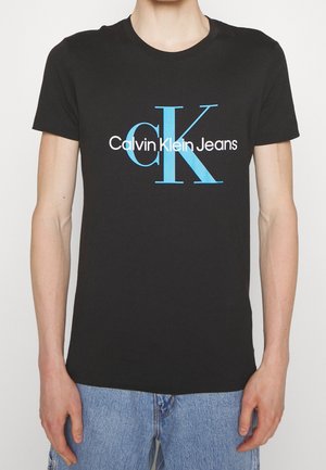 Personne portant un t-shirt noir avec un grand texte bleu "cK" et blanc "Calvin Klein Jeans", associé à un jean en denim bleu clair.