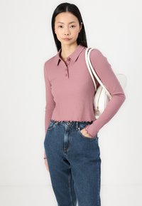 Ribbede langermede polo-skjorte i støvete rosa med krage, fem-knappers placket og volangkant; kombinert med høyt midje blå jeans.