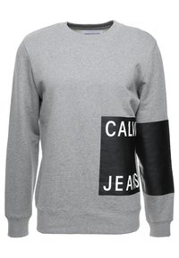 Šedá mikina z bavlněné směsi, která má černou umělou koženou část s bílým textem "CALVIN KLEIN JEANS" na přední straně. Klasický střih s kulatým výstřihem.