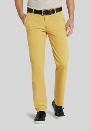 Mann trägt senfgelbe Hose, schwarzen Gürtel und schwarze Sneakers und steht vor grauem Hintergrund.