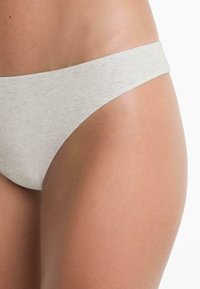 Close-up van de heup en bovenbeen van een persoon die lichtgrijze geribbelde onderkleding draagt, op een witte achtergrond.