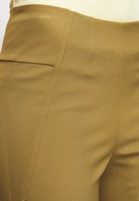 Vysoké kalhoty s přiléhavým střihem v tlumené hořčicové barvě, s hladkým povrchem a jemným prošíváním podél švů.