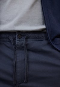 Gros plan sur un pantalon bleu marine à taille élastique, bouton et cordons, porté avec une chemise gris clair et une veste bleu marine.