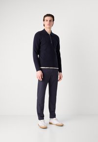 Marinblå långärmad polo med dragkedja vid kragen, kombinerad med mörka byxor och vita sneakers. Slät textur, åtsittande design och neutrala accenter.