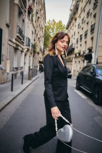 Mustas ülikonnas naine, kellel on pikad kõrvarõngad, kõnnib linnatänaval, hoides hõbedast süda-kujulist kotikest pärlkäepidemega.